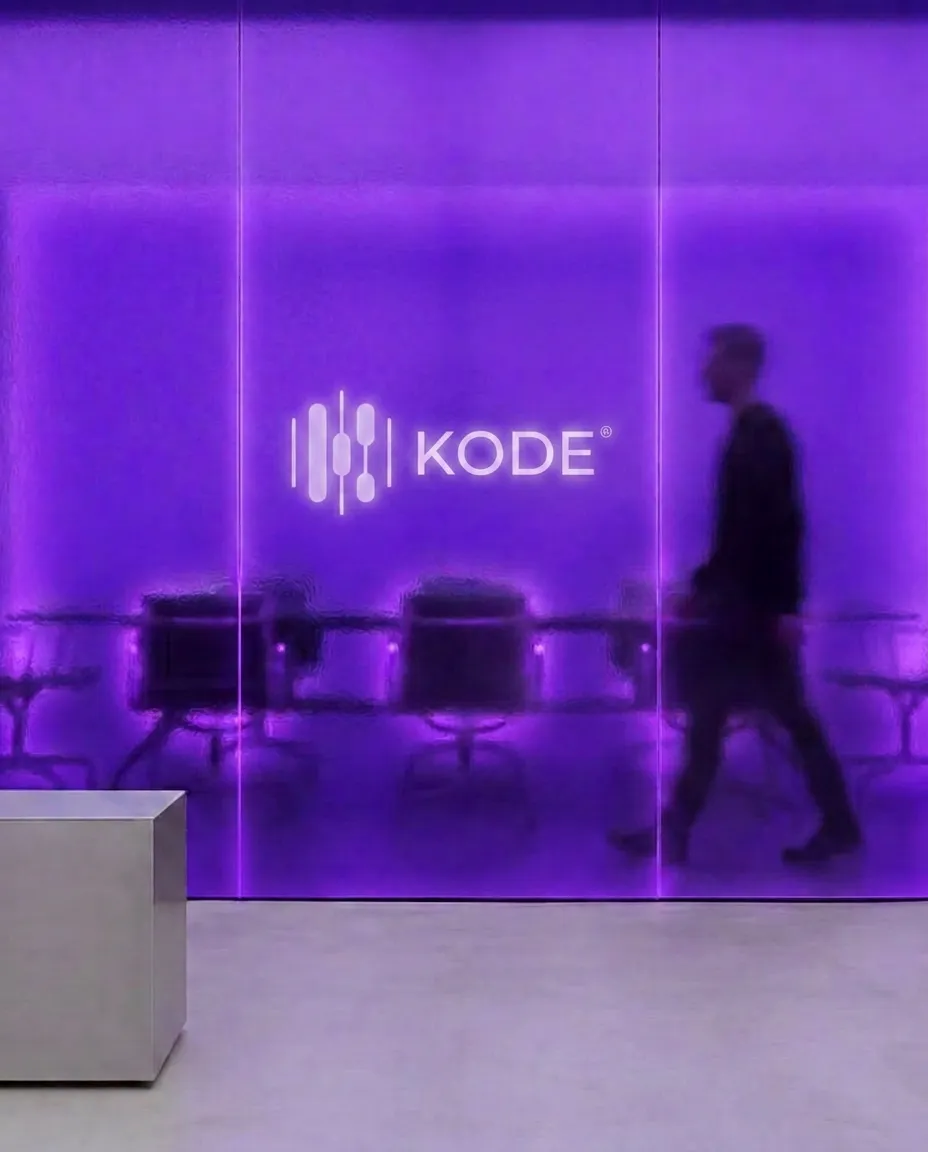 Person geht an einer violett beleuchteten Glaswand mit KODE® Logo vorbei, dahinter ein leerer Besprechungsraum